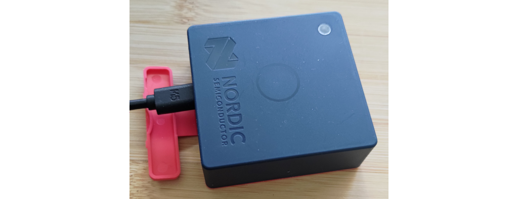 Nordic Semiconductor Thingy:53, 6cm grey box with Nordic logo, button, and light, with an open red flag and USB-C cord inserted