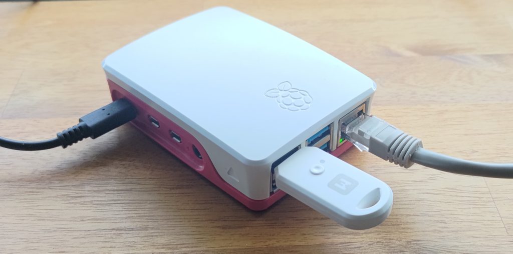 MakerDiary Thread Dongle in Raspberry Pi, the red and white Pi on a desk, showing the various ports, with USB-C power and RJ-45 ethernet connections, and a USB dongle sticking out the front